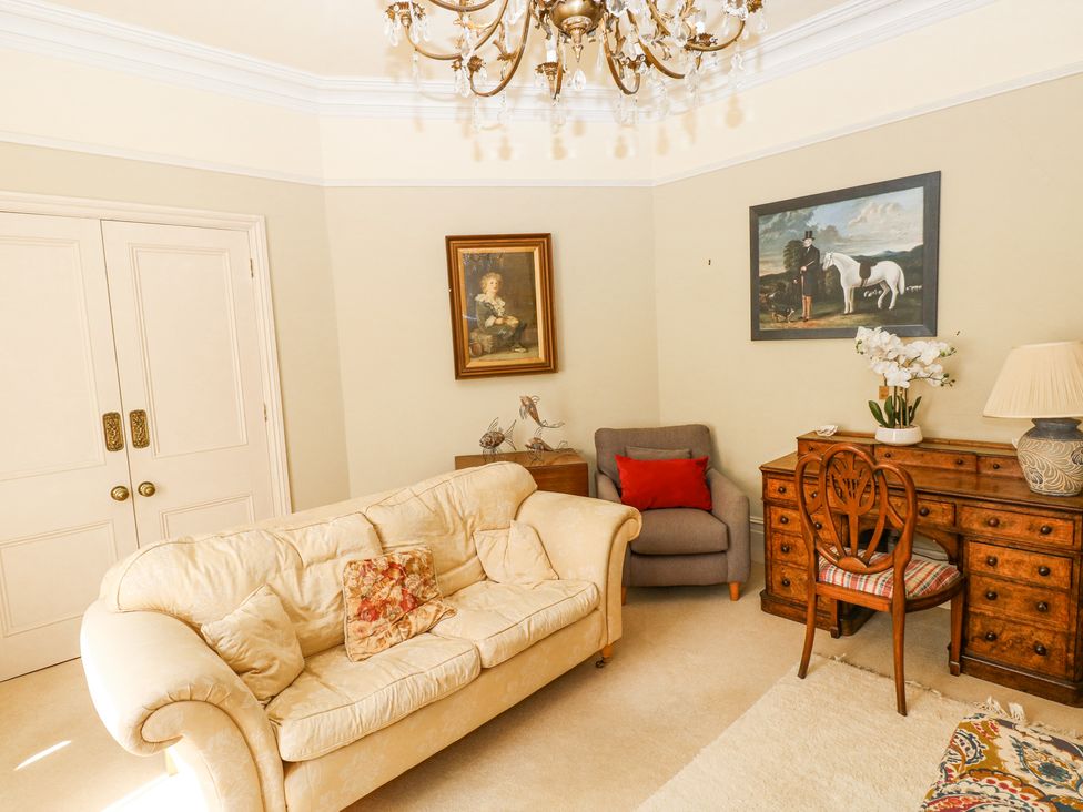 A sitting room with a cream sofa and floral cushions a gray armchair with a red cushion a wooden desk with a chair and lamp and two paintings on the walls at Hereford House in Tenby