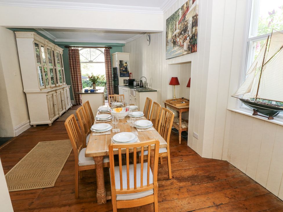 A dining room with a wooden dining table set for eight with plates and glasses a large cabinet an open kitchen area and a model sailboat on the windowsill at Hereford House in Tenby