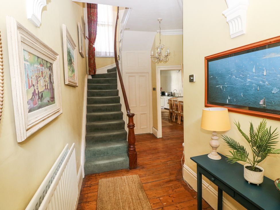 A hallway with wooden floor a green carpeted staircase framed artwork a dark green table lamp and a plant at Hereford House in Tenby