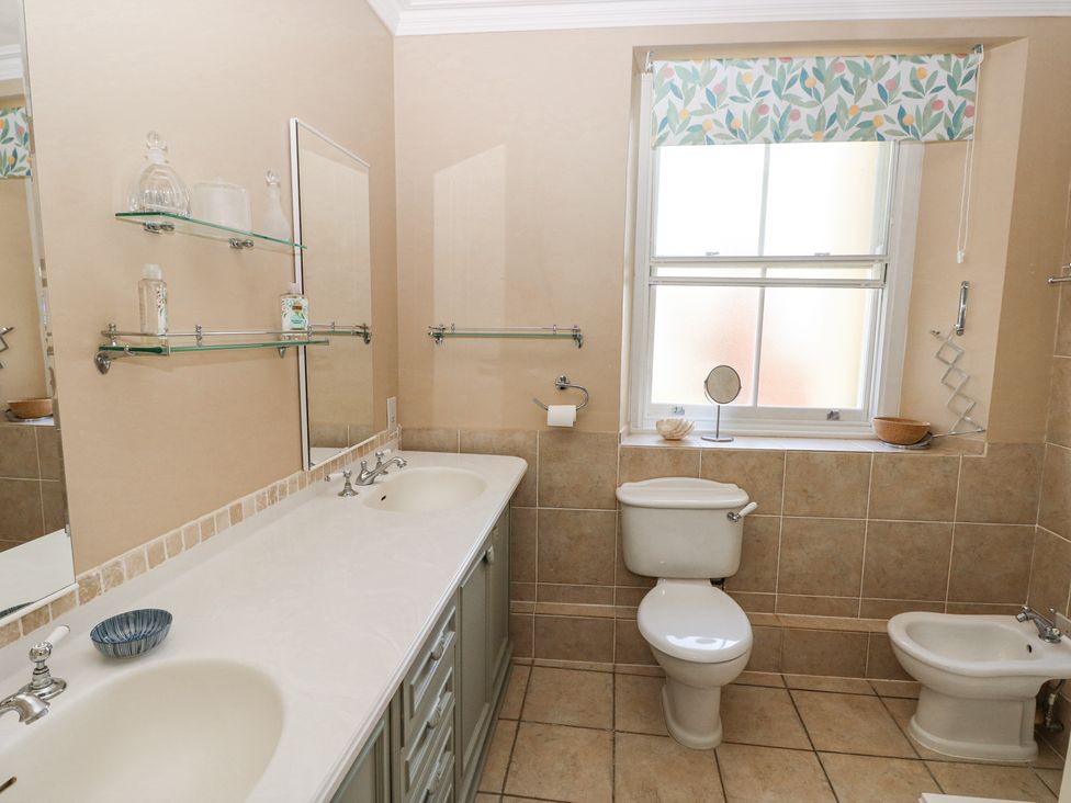 A bathroom with a double sink countertop toilet bidet window with floral valance shelves with bottles and small mirror