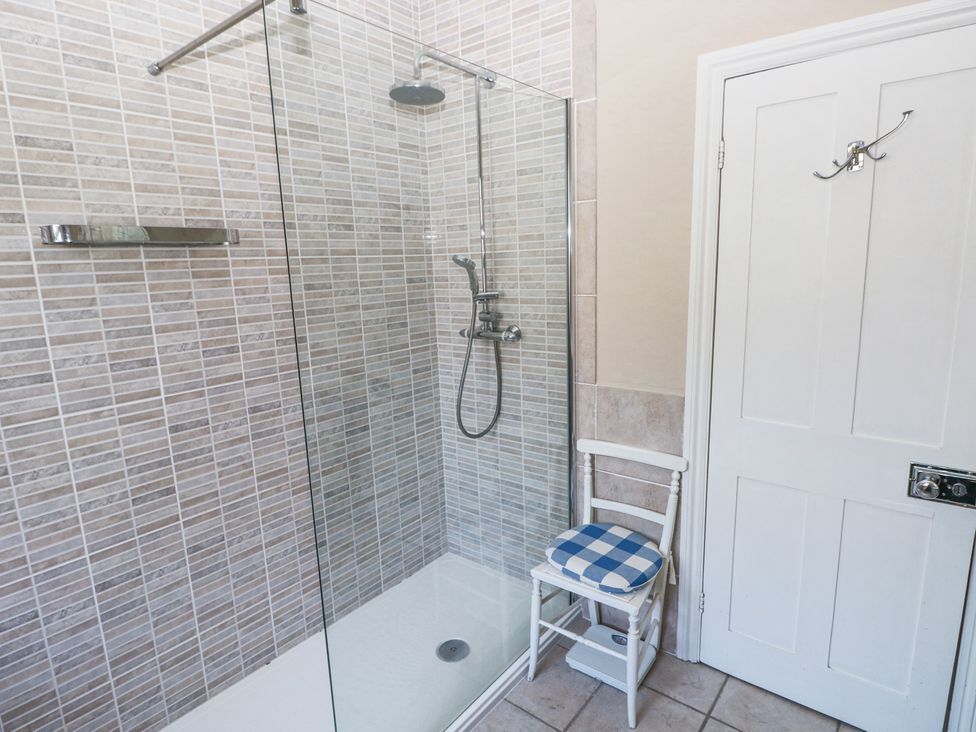 A bathroom with a glass shower enclosure tiled walls and a white chair with a blue checkered cushion near a white door