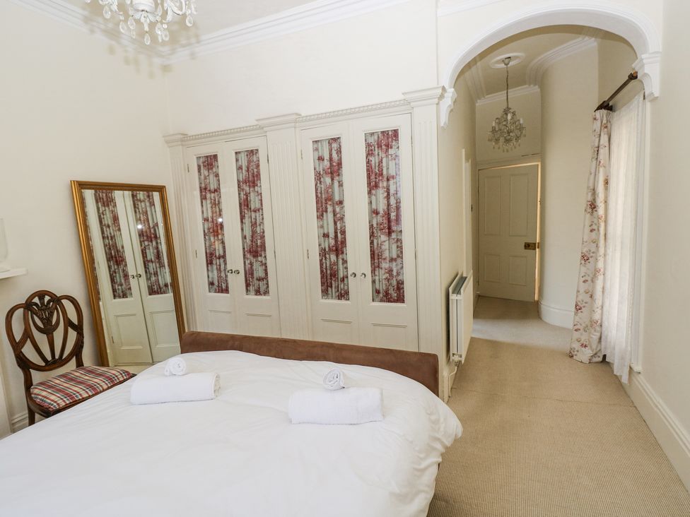 A bedroom with a bed and two rolled towels a wooden chair a full-length mirror built-in wardrobes with floral curtains an arched doorway and a window with curtains at Hereford House in Tenby