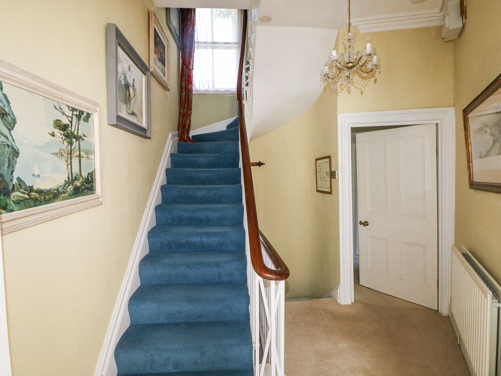 A staircase with blue carpet and paintings on the walls in a hallway with a chandelier and an open door at Hereford House in Tenby