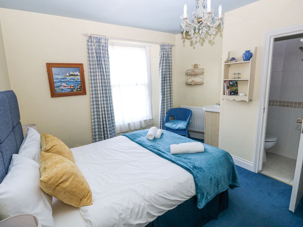 A bedroom with a bed with blue and yellow bedding a window with checkered curtains a blue chair a small sink and an en suite bathroom at Hereford House in Tenby