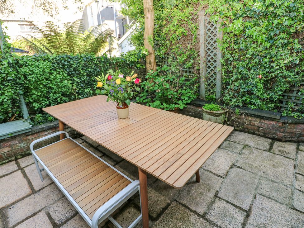 An outdoor patio with a wooden table and bench surrounded by green plants and flowers at Hereford House in Tenby