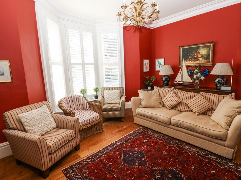 A living room with red walls, a beige sofa, striped chairs, a wicker chair, a patterned rug, lamps, and framed artwork at Hereford House in Tenby