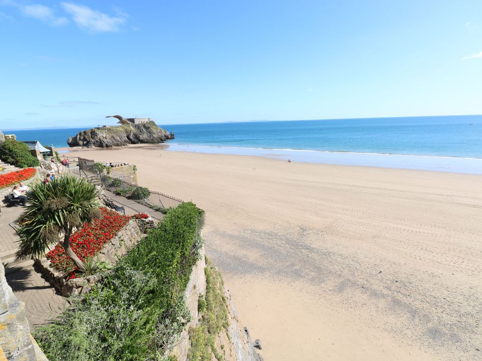 A wide sandy beach with a rocky island and a stone wall with red flowers and greenery overlooking the beach at Hereford House in Tenby