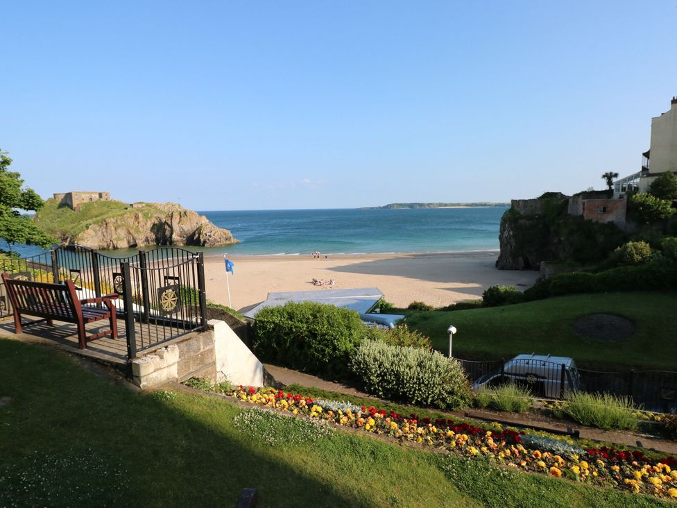 A view of a beach with a gate, bench, flowers, grassy area, and a rocky island in the sea at Hereford House in Tenby