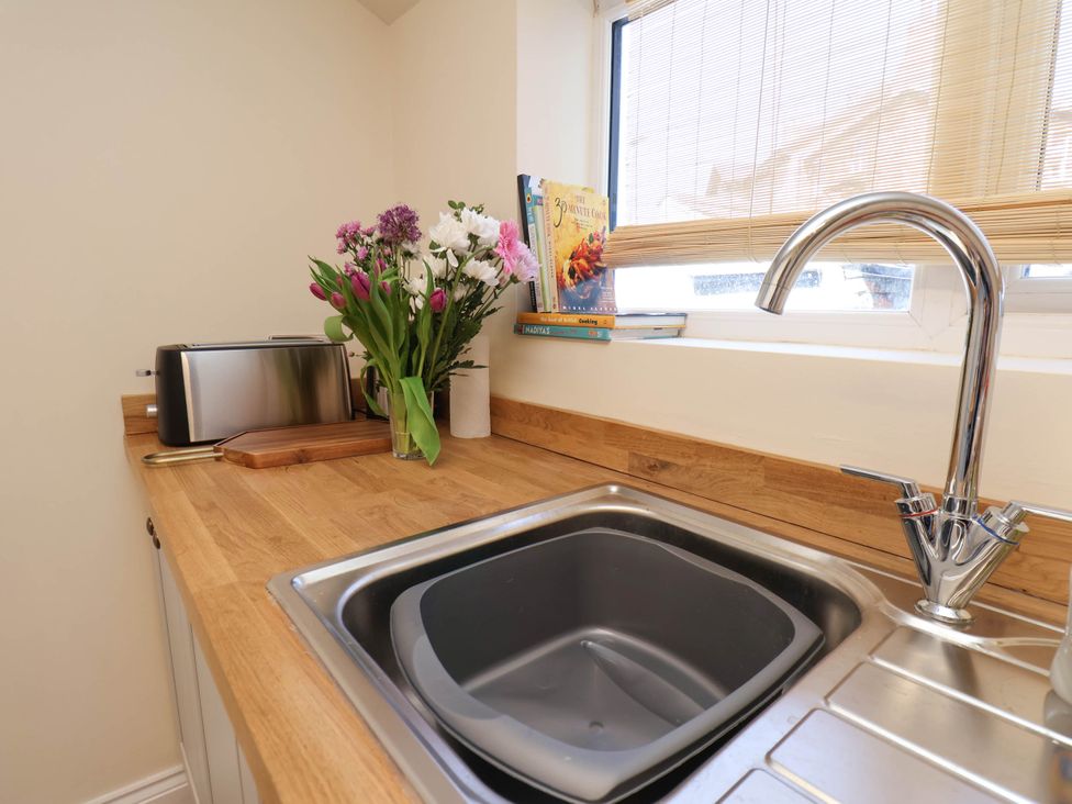A kitchen sink with a silver faucet a drying rack in the sink and a wooden countertop with a toaster flowers cutting board and cookbooks near the window at Flat 1 in Scarborough