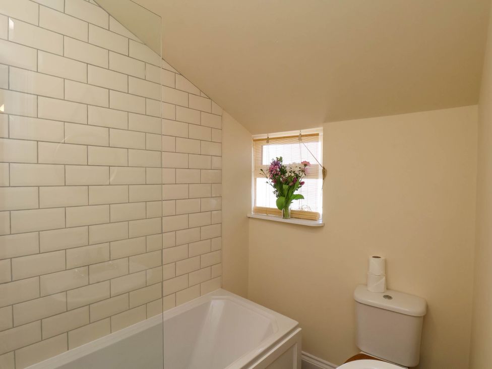 A bathroom with a bathtub, tiled wall, window with blinds and flowers, and a toilet with two rolls of toilet paper on top at Flat 1 in Scarborough