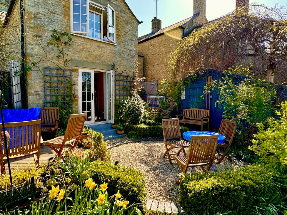 An outdoor garden area with wooden chairs around a blue round table and a wooden bench near a stone house and a blue shed