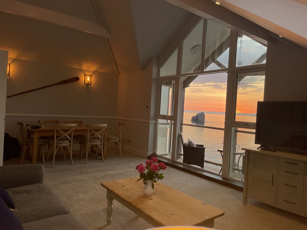 A living room with a wooden dining table and chairs a wooden coffee table with flowers a gray sofa and large windows showing a sunset over the sea at 12 Thurlestone Rock in Thurlestone