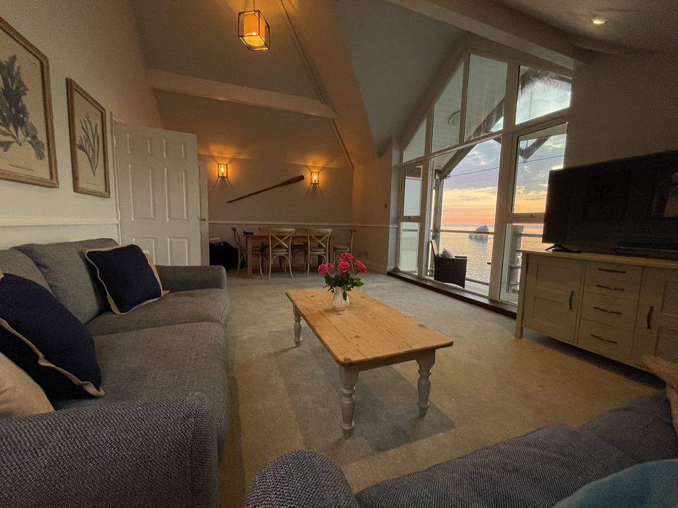 A living room with two sofas a wooden coffee table with flowers a dining table in the back and large windows overlooking water at 12 Thurlestone Rock in Thurlestone
