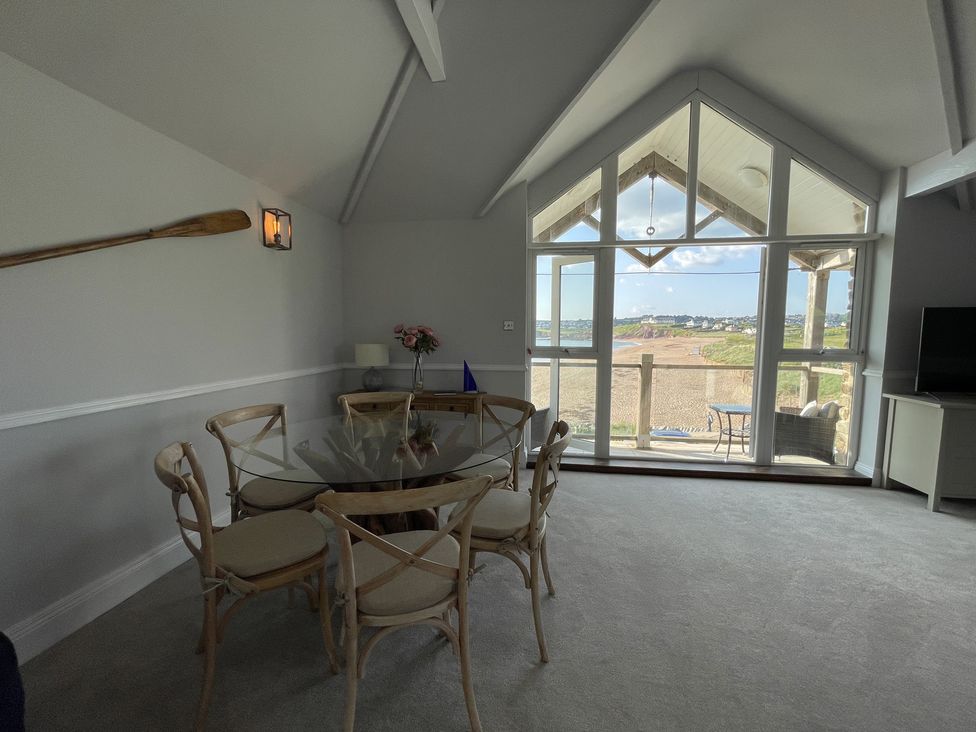 A dining area with a glass table and six chairs near large windows overlooking a beach at 12 Thurlestone Rock in Thurlestone