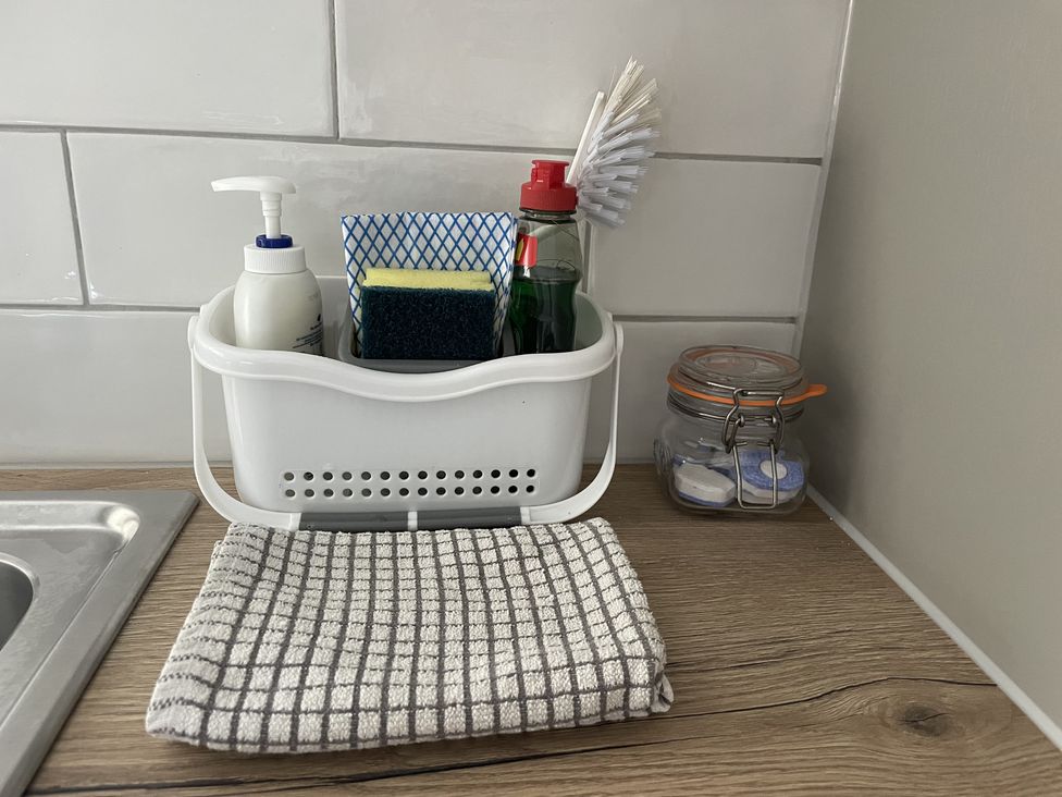 A kitchen countertop with a white caddy holding a soap dispenser sponge dish brush and a checkered cloth next to a glass jar with cleaning tablets at 12 Thurlestone Rock in Thurlestone