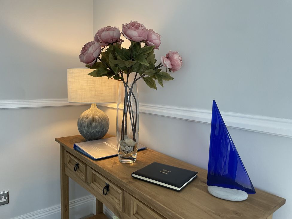 A wooden console table with a lamp a vase of flowers a guest book and a blue glass sailboat at 12 Thurlestone Rock in Thurlestone