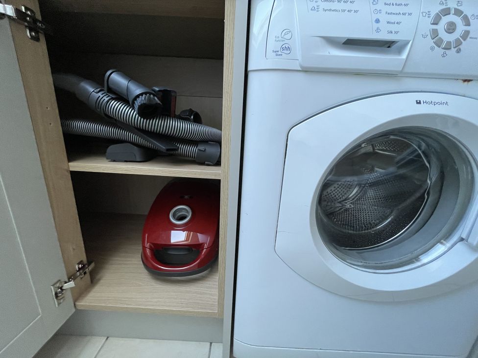 A washing machine next to an open cabinet with a red vacuum cleaner and hose inside at 12 Thurlestone Rock in Thurlestone