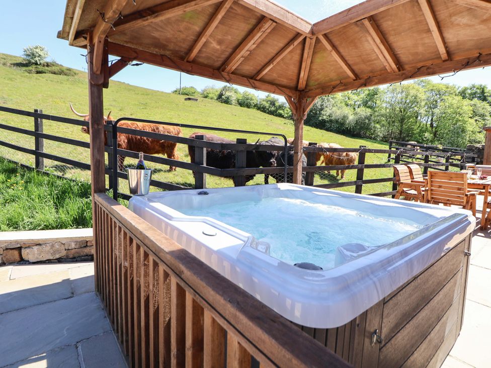 An outdoor hot tub under a wooden gazebo with a bottle in an ice bucket and wooden chairs and table near a fence with cows grazing at Parlour Barn in Leek