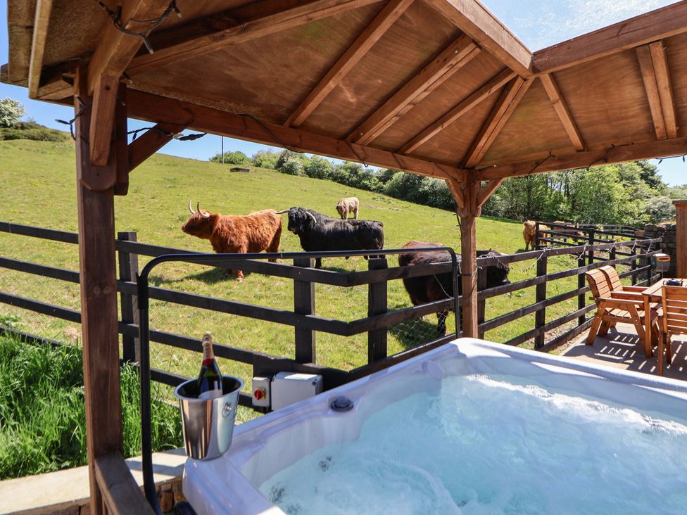 An outdoor hot tub under a wooden gazebo with a champagne bottle in an ice bucket and wooden chairs and table near a fenced grassy field with cows at Parlour Barn in Leek