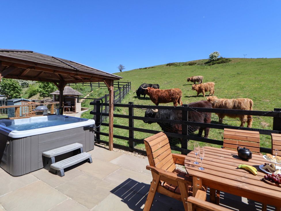 An outdoor patio with wooden table and chairs a hot tub with cover and a fenced grassy hill with cows at Parlour Barn in Leek