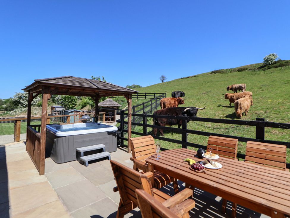 An outdoor patio area with a hot tub under a wooden gazebo and a wooden dining table with chairs next to a fenced grassy hill with cows at Parlour Barn in Leek