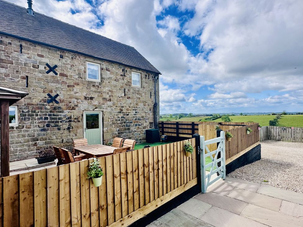A stone building with outdoor wooden table and chairs behind a wooden fence with a gate overlooking green fields at Pastures Barn Bradnop near Leek