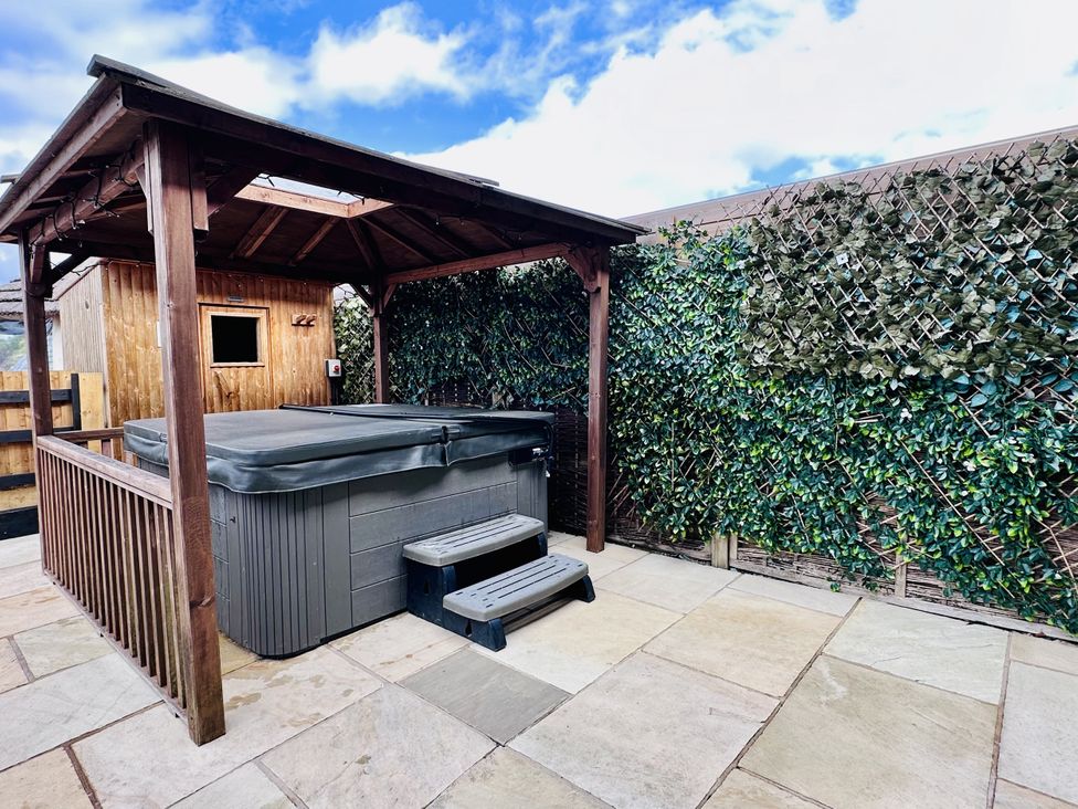 A covered hot tub with steps on a tiled patio next to a wooden shed and a hedge fence at Pastures Barn in Bradnop near Leek