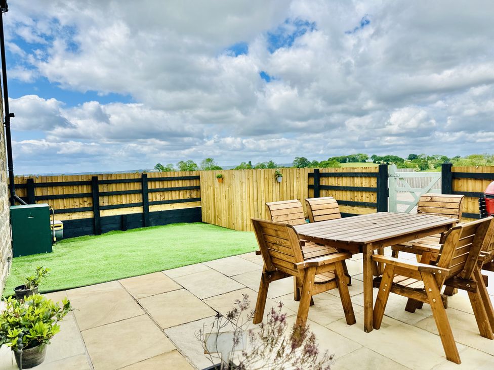 An outdoor patio with wooden table and chairs on a stone floor next to a grassy area enclosed with a wooden fence at Pastures Barn in Bradnop near Leek