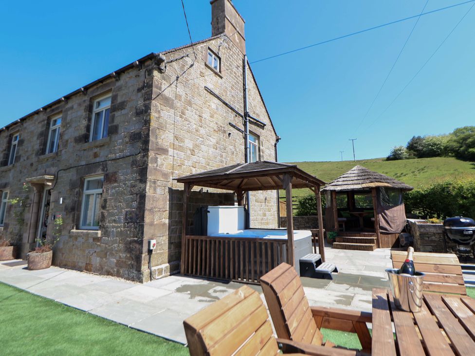 An outdoor patio area with a stone house a wooden hot tub cover a gazebo wooden chairs and table with a bucket and bottle at HollyDale Bradnop near Leek
