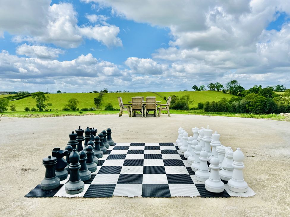 An outdoor concrete area with a large chess set on the ground and wooden chairs with a table in the distance at HollyDale Bradnop near Leek