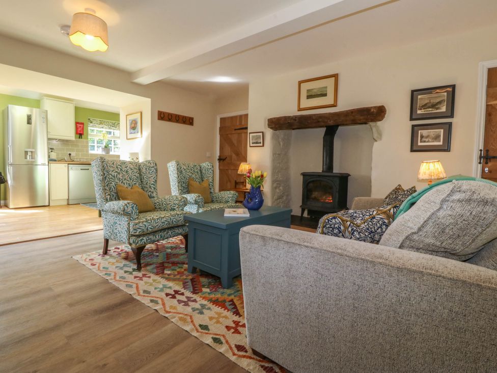A living room with patterned armchairs a blue coffee table and a wood stove adjacent to a kitchen area at Bodior Garden Cottage in Rhoscolyn