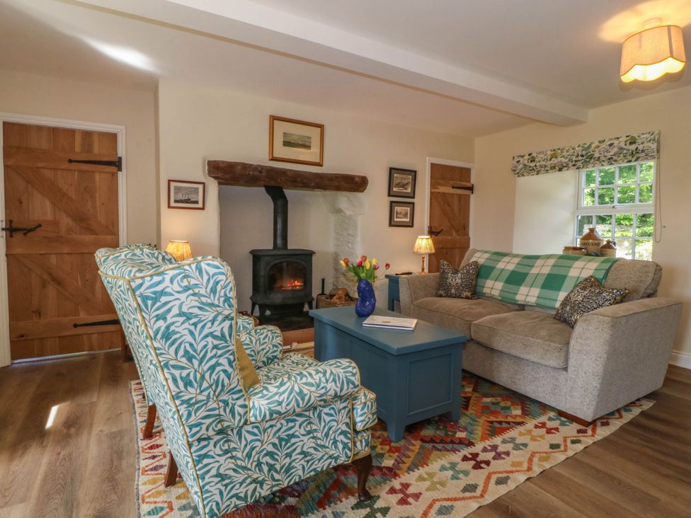 A living room with patterned armchairs a wood burning stove a sofa a coffee table and wooden doors at Bodior Garden Cottage in Rhoscolyn