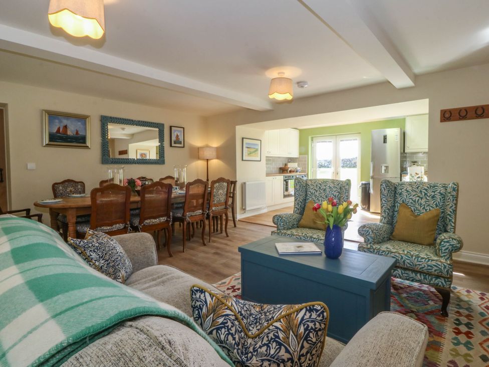 An open living and dining area with patterned armchairs a dining table and kitchen appliances at Bodior Garden Cottage in Rhoscolyn