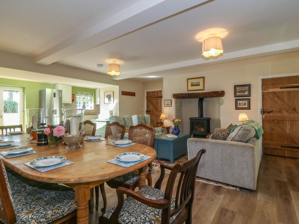 A combined dining and living room with a wooden dining table set with plates and chairs and a sitting area with sofas a stove and wooden doors at Bodior Garden Cottage in Rhoscolyn