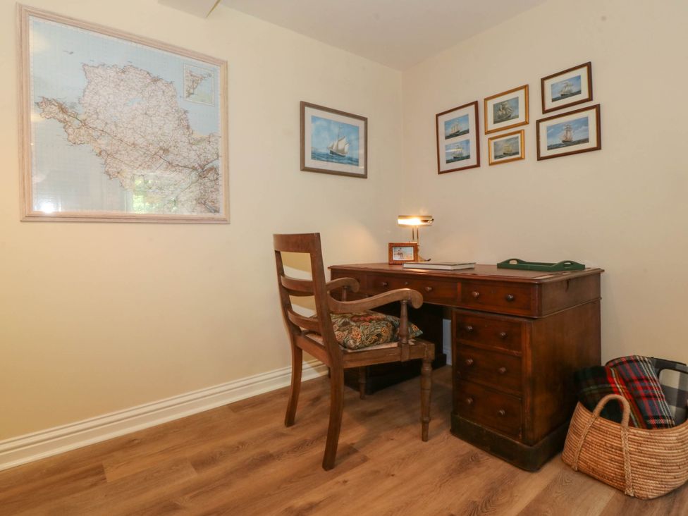 A wooden desk with a chair a lamp and framed pictures on the wall with a basket containing blankets on the floor at Bodior Garden Cottage in Rhoscolyn