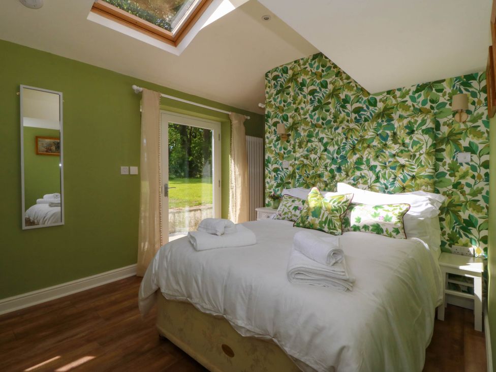 A bedroom with a bed with white linens and green leaf patterned pillows against a green leaf wallpaper wall next to a glass door and a vertical mirror on the green painted wall at Bodior Garden Cottage in Rhoscolyn