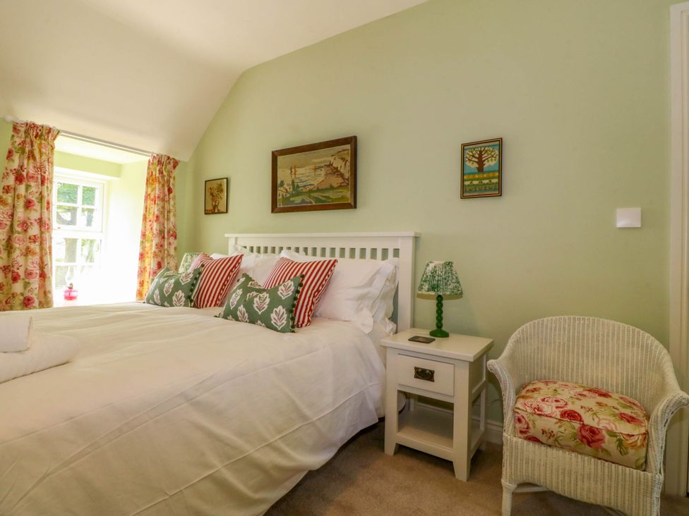 A bedroom with a double bed with striped and floral pillows a bedside table with a lamp a wicker chair with a floral cushion and floral curtains at Bodior Garden Cottage in Rhoscolyn