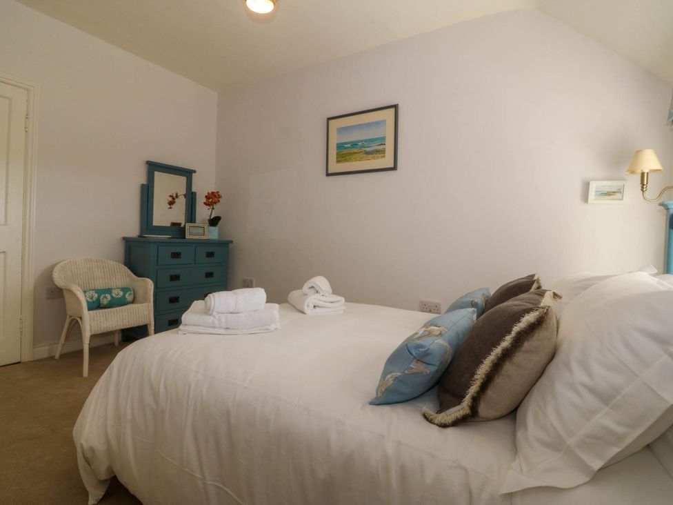 A bedroom with a bed, pillows, folded towels, a blue dresser with a mirror, a wicker chair, and framed pictures on the wall at Bodior Garden Cottage in Rhoscolyn