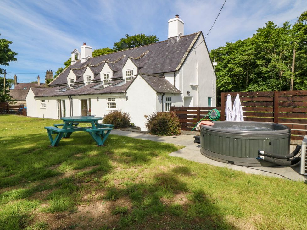 A white cottage with multiple windows and chimneys a green picnic table and a round hot tub in the garden at Bodior Garden Cottage in Rhoscolyn