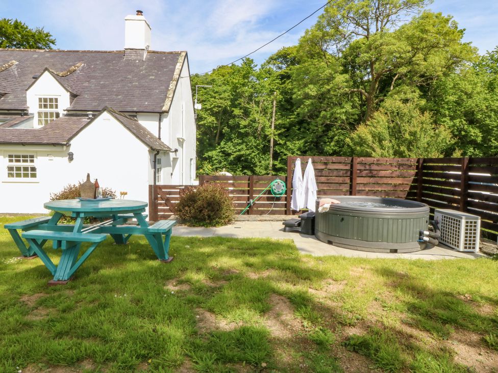 A garden with a green picnic table a hot tub a white house and trees at Bodior Garden Cottage in Rhoscolyn