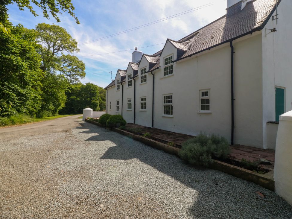 A white house with multiple windows and a gravel driveway next to a tree-lined road at Bodior Garden Cottage in Rhoscolyn