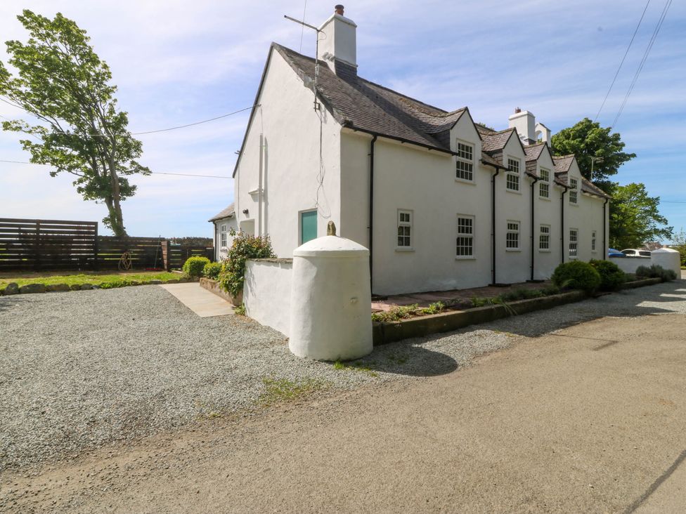 An exterior view of a white two-story house with multiple windows next to a gravel driveway and a fenced garden area at Bodior Garden Cottage in Rhoscolyn