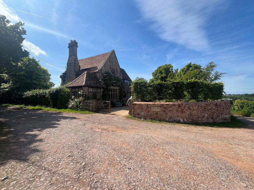 An old stone house with a chimney and a tiled roof behind a stone wall and wooden gate at The Gables in Spaxton near Nether Stowey