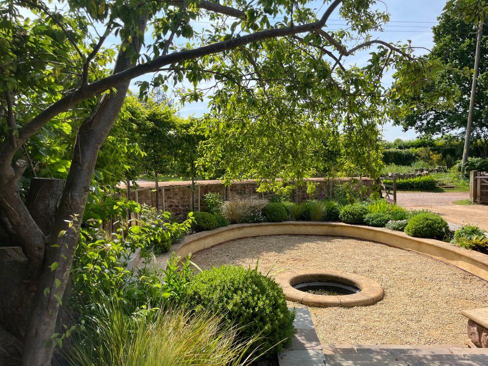 A garden with gravel ground and a circular stone water feature surrounded by greenery and a tree at The Gables in Spaxton near Nether Stowey