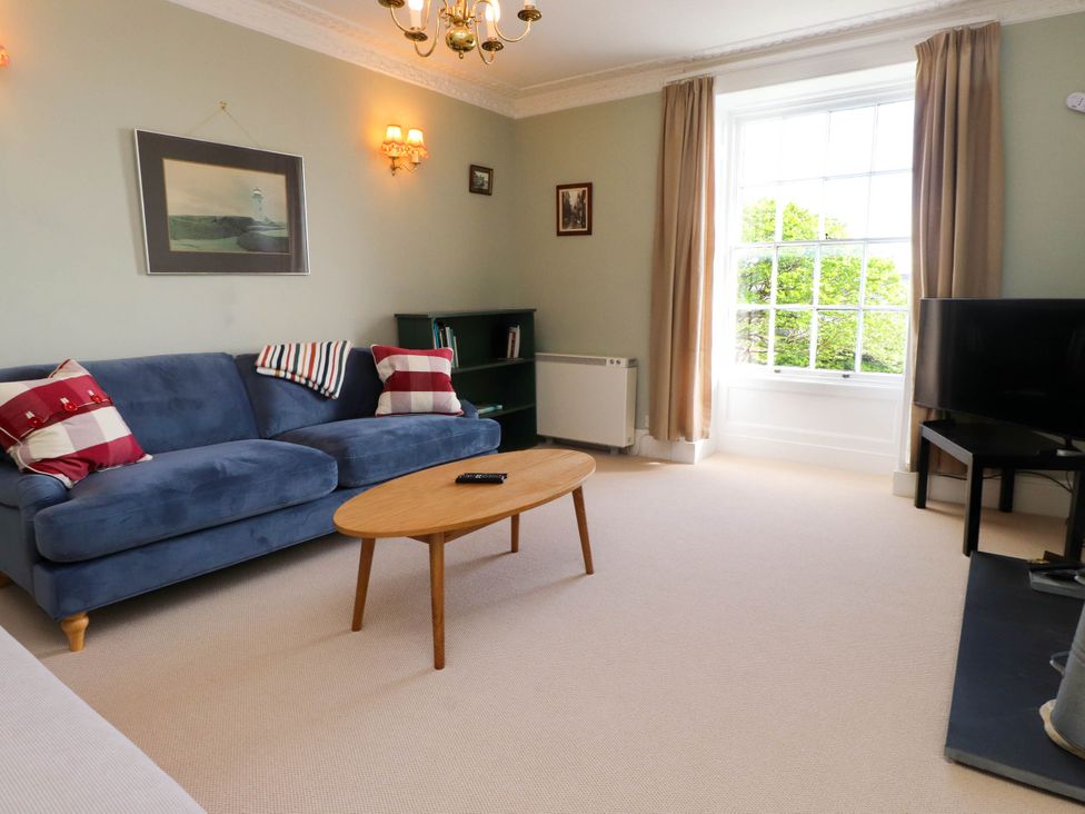 A living room with a blue sofa red and white pillows a wooden coffee table a television and a large window with curtains at Flat 3 in Trearddur Bay