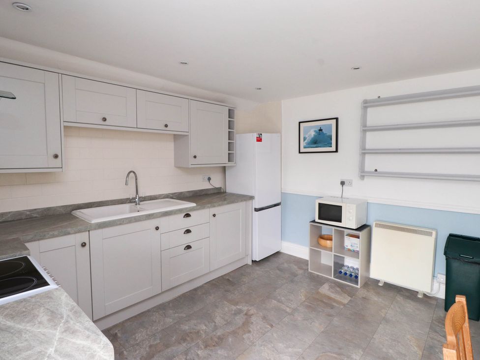 A kitchen with white cabinets a sink a refrigerator a microwave on a shelf and a trash bin at Flat 3 in Trearddur Bay
