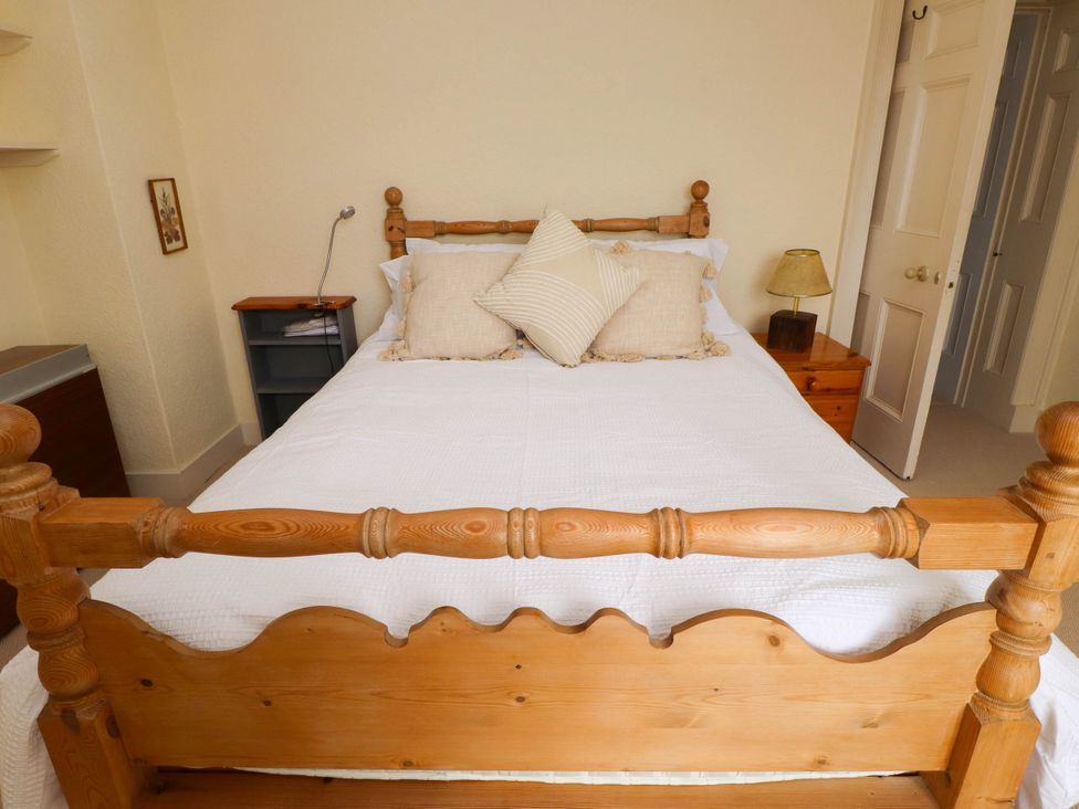 A bedroom with a wooden bed with white bedding beige pillows a bedside table with a lamp and an open door at Flat 3 Trearddur Bay