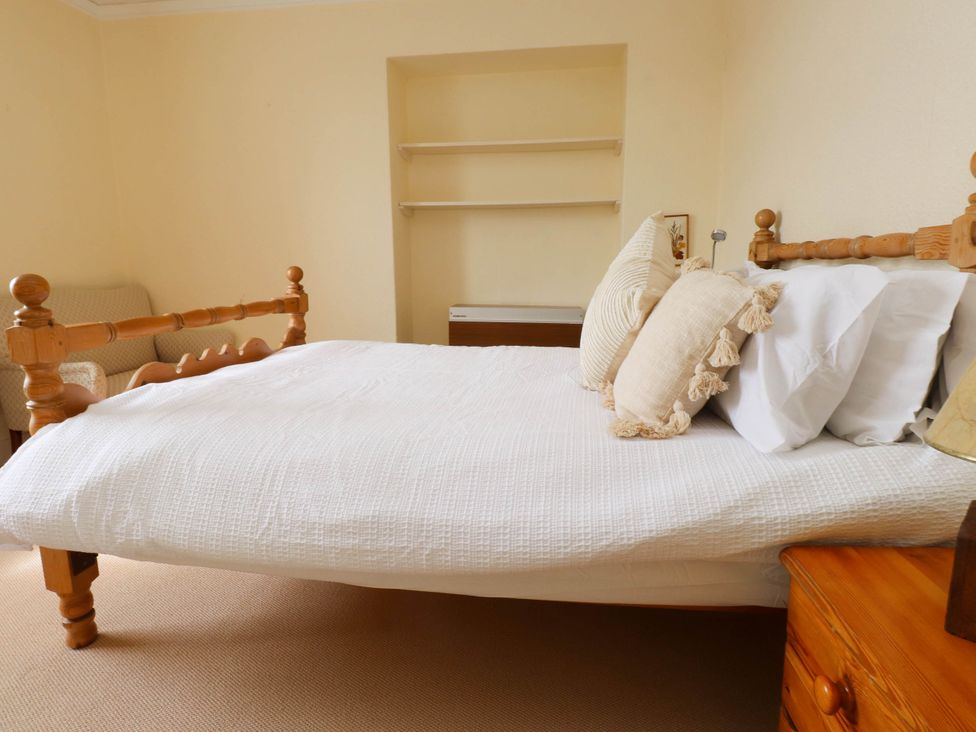 A bedroom with a wooden bed and white bedding next to a wooden nightstand and beige armchair at Flat 3 in Trearddur Bay