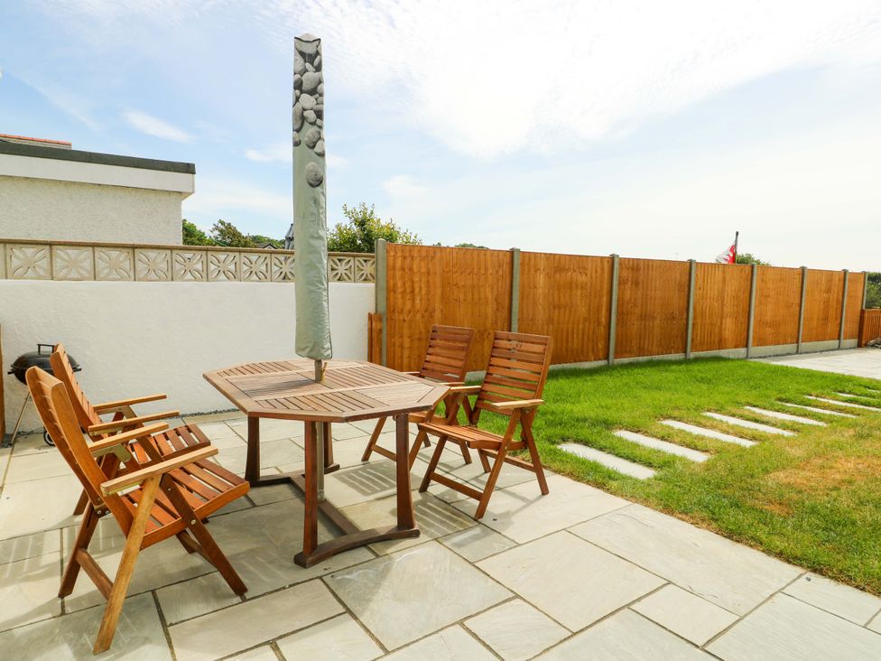 A patio with a wooden table and four wooden chairs next to a grass lawn and wooden fence at Ty Bach in Llanfaelog near Rhosneigr