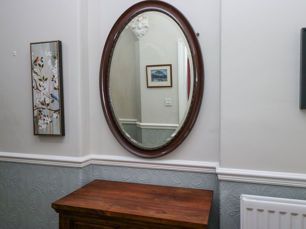An oval mirror above a wooden cabinet with a framed picture and light switch reflected on the wall at Sunnycroft in Keswick